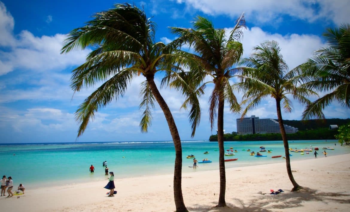 man in black shirt sitting on white sand near palm tree during daytime